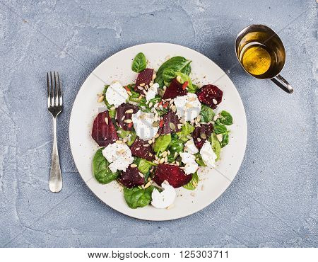 Salad with roasted beetroot, spinach, soft goat heese and seeds in light plate over grey concrete textured background, oil in saucer. Top view