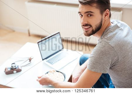 Smiling handsome young man using laptop and eating cerials with milk at home