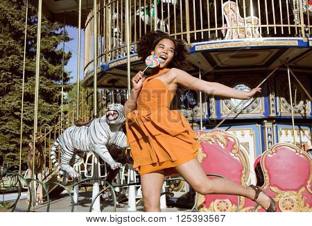 cool real modern teenage girl with candy near carousels at amusement park walking, having fun