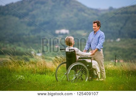 Senior man with woman sitting in wheelchair, holding hands, oustide in green autumn nature, rear view