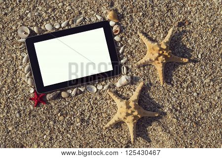 Photo frame made of seashells and pebbles with two starfish placed next to it and tablet computer within the frame with blank screen on the beach
