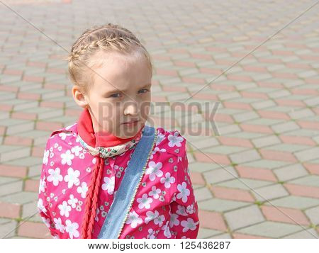 Portrait of small pensive Caucasian schoolgirl in a pink jacket on the square paved with colored tiles