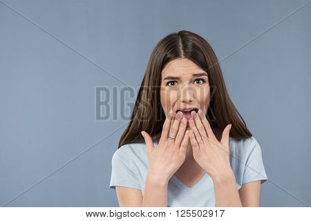 I am afraid. Pleasant young scared girl holding her hands in front of her face and feeling horrified while standing isolated on grey background