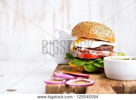 Breakfast set. Homemade beef burger with fried egg and vegetables, onion rings and coffee cups on wooden board, white painted background. Selective focus, copy space