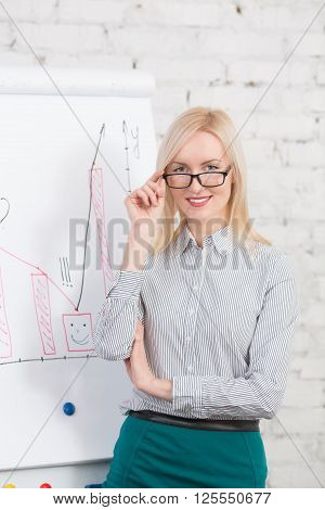 Businesswoman giving explication in front of growing chart on whiteboard during meeting in office. Happy lady in glasses smiling.
