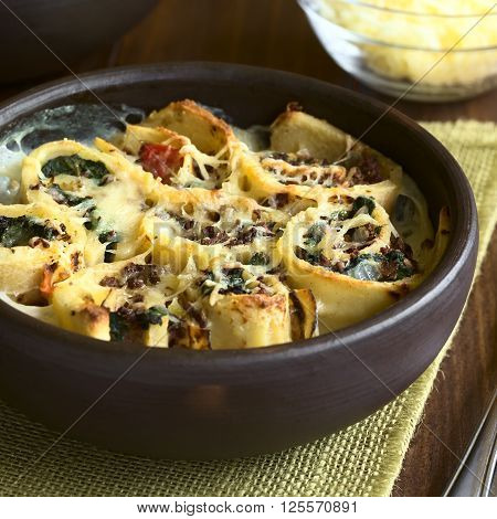 Baked crepe rolls casserole filled with spinach tomato and mincemeat with cream sauce and grated cheese on top served in rustic bowl photographed with natural light (Selective Focus Focus on the front of the middle roll)