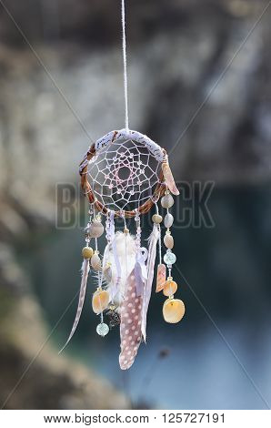 Handmade pink native american dream catcher on background of rocks and lake. Tribal elements, feathers, shells, lace