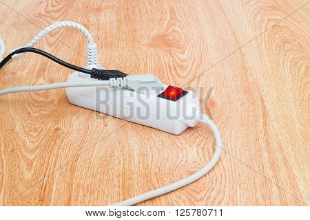 White power strip with three electrical sockets and illuminated power circuit breaker and three different power plugs with mains cables on a wooden surface