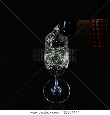 Pouring process of sparkling wine into champagne glass from champagne bottle splashes drops and froth around glass against black background