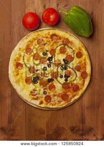 A photo of a pepperoni pizza with salami and peppers with shiny red tomatoes and a green bell pepper on a pizza stone on a dark wooden background with a place for text
