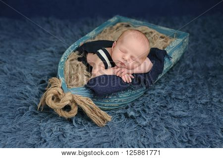 Portrait of a two week old newborn baby boy. He is sleeping in a miniature boat and wearing dark blue overalls. Shot in the studio on a blue flokati rug.