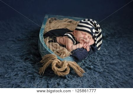 Portrait of a two week old newborn baby boy. He is sleeping in a miniature boat and wearing a dark blue and white striped hat and overalls. Shot in the studio on a blue flokati rug.