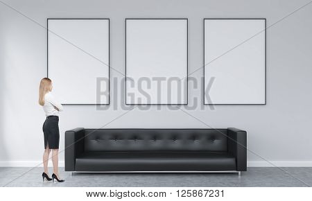 Three white picture frames above black sofa in concrete interior with businesswoman lookign at them. Mock up 3D Rendering