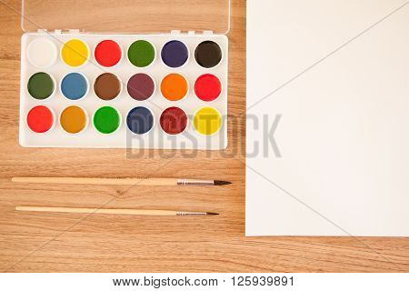 Artist's workspace. Set of 18 watercolor paints in white base sheet of white watercolor paper 2 brushes on light woden background with copyspace. Artistic background.