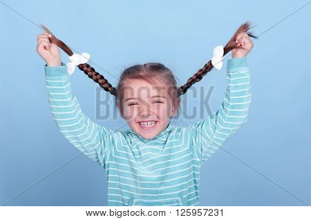 Laughing kid girl 3-4 year old having fun over blue. Wearing blue striped hoodie. Holding braids. Smiling baby.