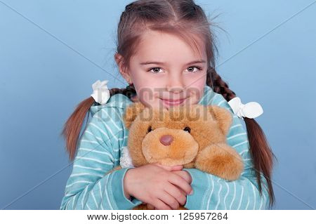 Smiling kid girl 3-4 year old playing with teddy bear over blue. Wearing blue striped hoodie.
