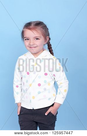 Smiling kid girl 3-4 year old with hands in pockets posing over blue. Wearing hoodie and pants. Childhood. Stylish.