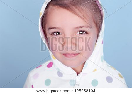 Cute baby girl 3-4 year old posing in room over blue. Wearing white hoodie with colorful polka dots. Looking at camera. Closeup portrait.