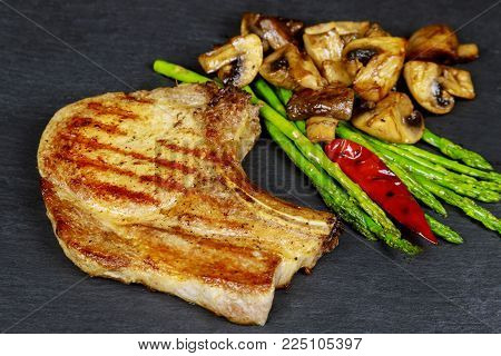 Grilled pork steaks with herbs, asparagus, mushrooms on stone slate background close up. Hot Meat Dishes. Top view