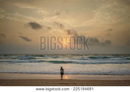 Woman traveler standing in the distance on the beach and looking at the sunset. View from the back