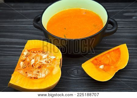 Pumpkin cream soup in a black ceramic bowl and pieces of fresh pumpkin on black wooden table