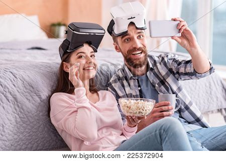One more shot. Awesome loving gay couple in VR glasses chortling while taking selfie and enjoying popcorn and coffee