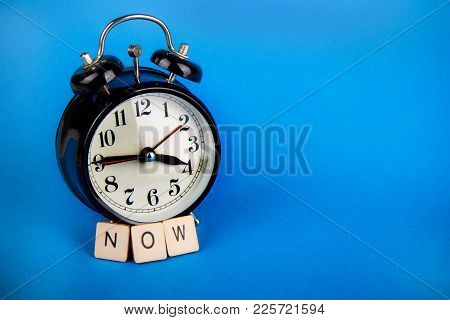 Black Alarm Clock With A Blue Background Showing Time With A Sign Saying Now