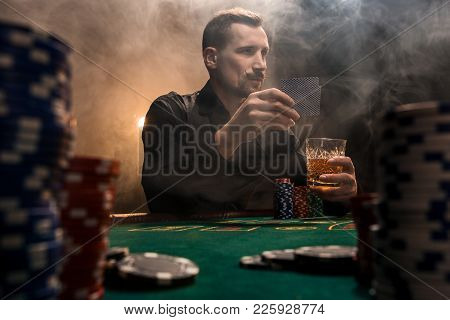 Young Handsome Man Sitting Behind Poker Table With Cards And Chips. In The Foreground Stacks Of Chip