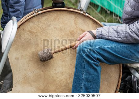 Big Drum And Drummer Sitting Next To Him With A Stick Outside. Street Musician With Street Clothes