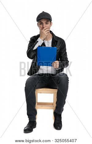 young casual man wearing a black leather jacket and hat sitting and holding his blue clipboard and toucing his face shocked against white studio background
