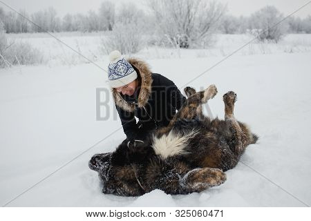 Cheerful Young Woman Embracing With Caucasian Shepherd Dog On Snow-covered Field In Frosty Winter Da