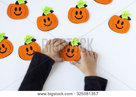 Top View Of Halloween Crafts, Felt Handmade Pumpkins On White Paper Background In The Hands Of A Chi