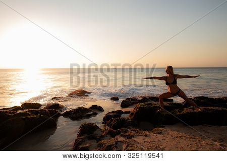Yoga Concept. Young Woman Practicing Yoga, Standing In Virabhadrasana Pose On The Beach. Silhouette 