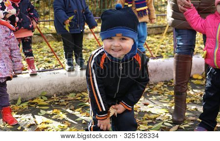 Autumn Time. Little Smiling Child With Yeallow Leaf Dressed In Black Jacket And Hat In The Kindergar