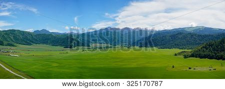 Mountain View With Sibirian Altai Mountains And Green Grass Field, Small Forest And Village, Blue Sk
