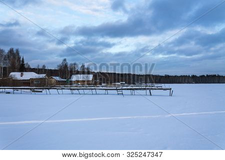Old Wooden Pier In The Snow. Wooden Shacks By A Frozen Lake. Pine Forest