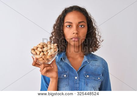 Young brazilian woman holding bowl with peanuts standing over isolated white background with a confident expression on smart face thinking serious