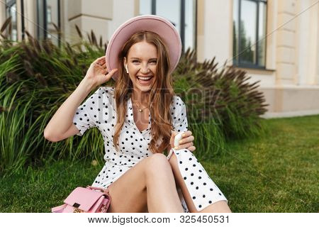 Image of amazing positive optimistic emotional cheery redhead woman sit on green grass outdoors listening music with earphones.
