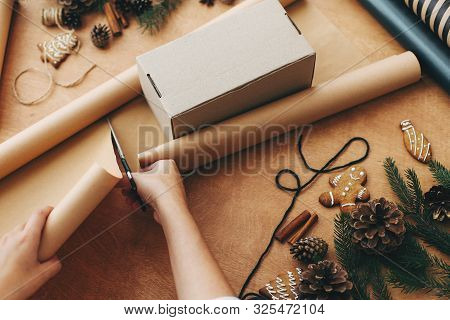 Hands Cutting Wrapping Paper With Scissors And Gift Box, Pine Branches, Cones, Gingerbread Cookies,
