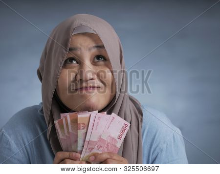 Portrait Of Indonesian Muslim Woman Holding Rupiah Money, Smiling And Thinking Gesture