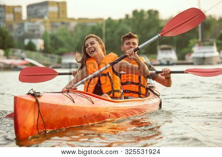 Happy Young Caucasian Couple Kayaking On River With Sunset In The Backgrounds. Having Fun In Leisure