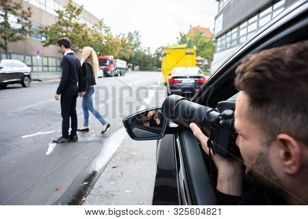 Young Male Spy Sitting In The Car Taking Photograph Of A Couple Walking On Street