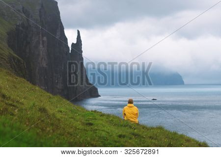 Tourist in yellow jacket looks at Witches Finger cliffs from Trollkonufingur viewpoint. Vagar island, Faroe Islands, Denmark.