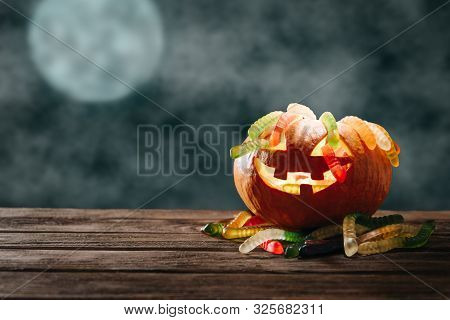 Glowing Carved Pumpkin With Colorful Candies In Form Of Worms On Wooden Table In Halloween Night.