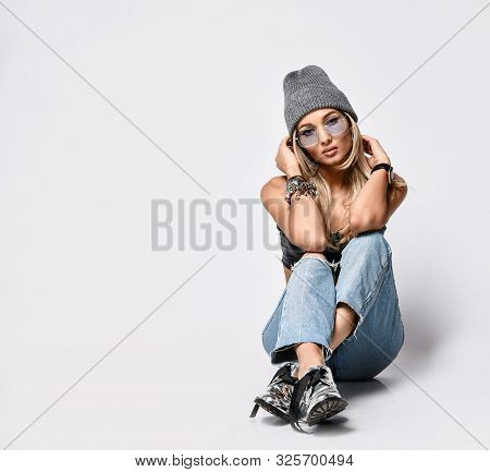 Sensual Grunge Style Blonde Woman In Blue Torn Jeans, Leather Bralet, Knitted Winter Hat And Glasses