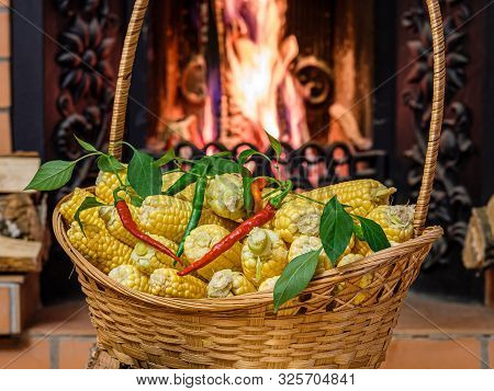 A Wicker Basket To The Top Filled With Ears Of Ripe Corn And Hot Peppers. In The Background A Burnin