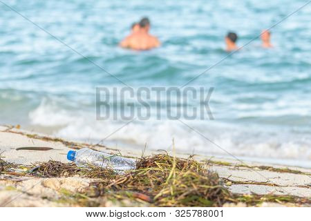 Close Up Image Of Empty Plastic Water Bottle On Dirty Beach Filled With Seaweed, Garbage And Waste O