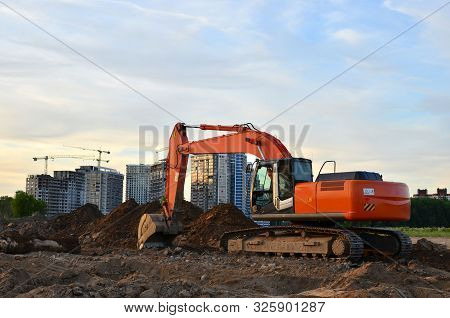 Crawler Excavator In Construction Site On The Background Of Tower Cranes And Multi-storey Residentia