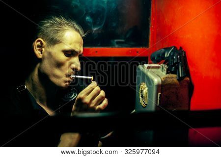 A Man Of Criminal Appearance In A Payphone Booth At Night. Talking On The Phone With A Revolver And