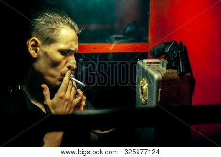 A Man Of Criminal Appearance In A Payphone Booth At Night. Talking On The Phone With A Revolver And